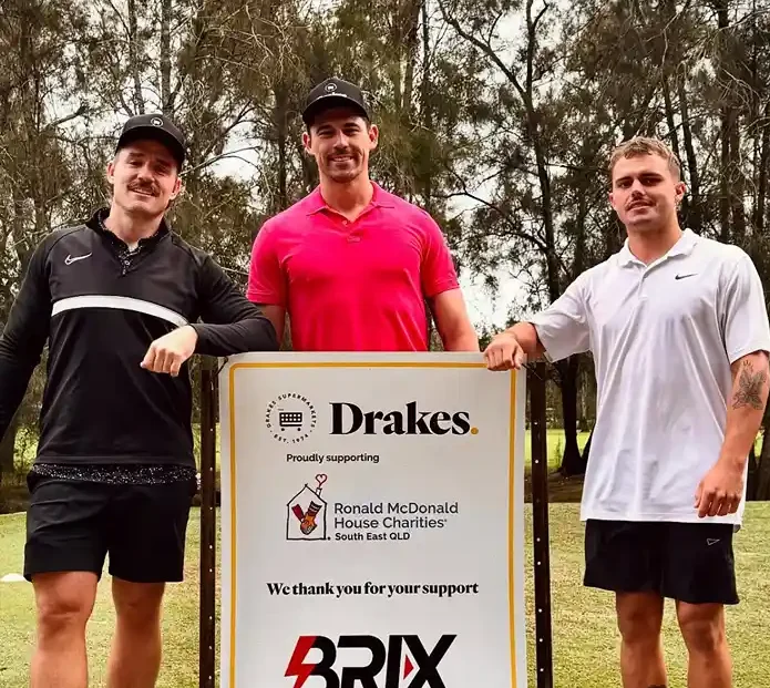 Three men standing beside a sponsorship sign supporting a charity, community support event.
