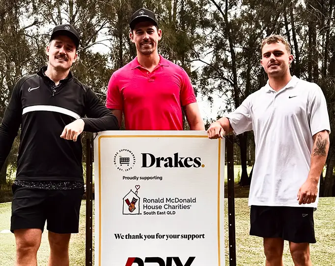 Three men standing beside a sponsorship sign supporting a charity, community support event.