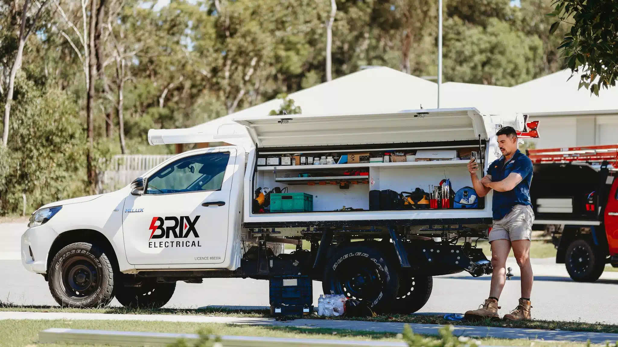 A white BRIX Electrical utility truck is parked on a residential street with its side service canopy open, revealing neatly organized shelves stocked with tools, equipment, and supplies.